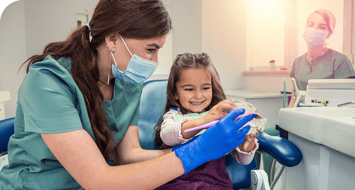 Em um consultório odontológico, uma dentista de jaleco verde, máscara e luvas azuis demonstra técnicas de escovação para uma menina sorridente, utilizando um molde de boca. A criança está sentada na cadeira do consultório, segurando a escova e interagindo com a profissional. Ao fundo, outra dentista acompanha a cena, também de máscara e jaleco verde, com luz natural entrando pela janela.